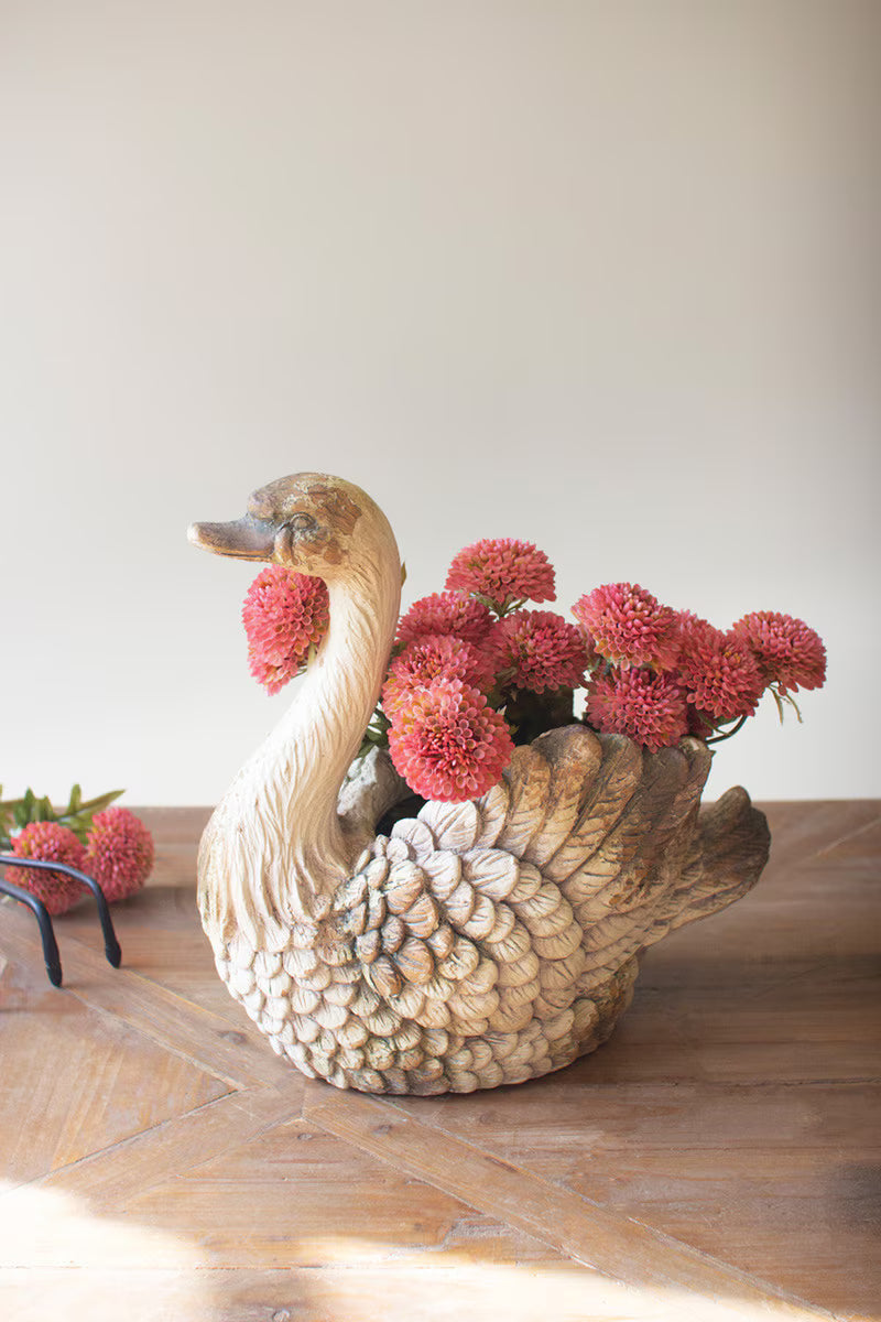 A decorative swan-shaped planter with a textured beige body, filled with red flowers, placed on a wooden table against a plain light background.